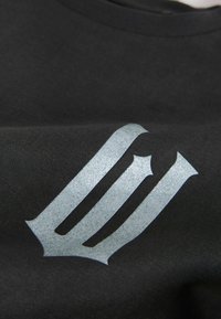 Must T-särk, millel on tekstuuriga hõbedane logo. Kangas on sileda tekstuuriga ja logol on stiliseeritud mitme triibuga muster.