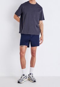 Dark gray cotton T-shirt with a chest pocket, paired with navy shorts. Model wears white socks and gray/white sneakers. Simple design.
