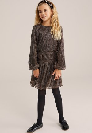 Schimmerndes braunes Plissee-Kleid mit langen Ärmeln, einer taillierten Taille und einem gestuften Rock. Schwarze Strumpfhosen und Schuhe runden das Outfit ab.