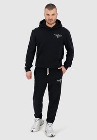 Zwarte hoodie en joggers van een katoenmengsel met een voorkeurszak en een trekkoord. Geborduurd logo detail. Witte sneakers maken de outfit compleet.