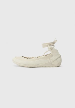Off-white flad sko med gummisål, ankel-remme og krydslægning på overdelen, vist fra siden på en ensfarvet baggrund.