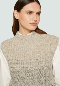 Gebreide sweatervest in beige met een textuurpatroon, voorzien van een hoge hals en brede armsgaten, gelaagd over een wit shirt.
