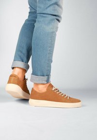 Zapatillas de ante marrón con suelas de goma beige, diseño de cordones y detalle del logo en el talón. Llevadas con jeans claros azules arremangados.