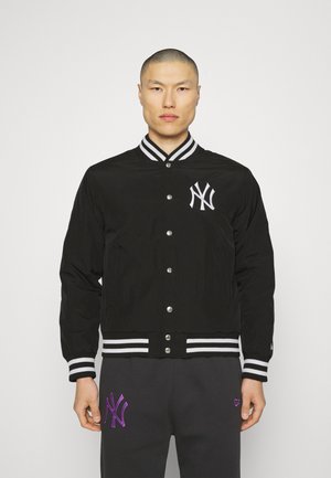 Chaqueta bomber negra con cuello y puños de rayas blancas, presenta un logo bordado blanco en el pecho y botones de presión plateados.