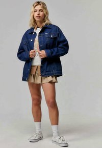 Denimjakke i mørkeblå med frontlommer, oversized passform, knappestolpe, lagd over en grafisk t-skjorte og beige skjørt. Hvite sneakers.