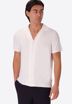 Uomo che indossa una camicia bianca a maniche corte abbottonata e pantaloni neri, in piedi con una mano in tasca e guarda di lato.