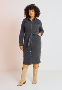 Vestido midi de mezclilla en gris oscuro, con mangas largas, botones, dos bolsillos en el pecho y un cinturón de lazo. Combinado con botas de cowboy bordadas en beige.