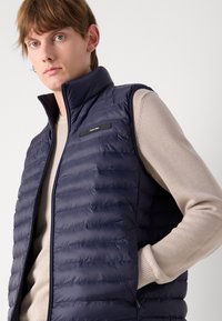 Jūras spēku pufīgs veste ar kvalificētu dizainu un stāvošu apkakli, ar priekšējo rāvējslēdzēju, sānu kabatām un logo akcents. Nēsāta pāri pelēkam džemperim.