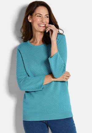 Vrouw die glimlacht en naar de zijkant kijkt, gekleed in een structuurtrui in turquoise met driekwartmouwen en donkerblauwe spijkerbroek.