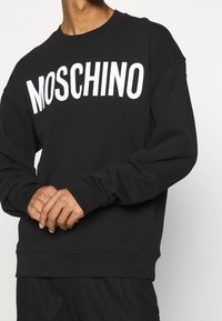 Sweatshirt noir en mélange de coton, avec le texte "MOSCHINO" blanc sur la poitrine, poignets et ourlet côtelés, coupe décontractée.