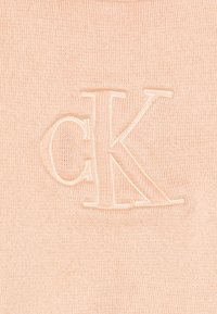 Tessuto lavorato a maglia color pesco chiaro con una sottile texture verticale. Presenta un logo "CK" embossato in un colore abbinato al centro.