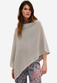 Beige neuleponcho, jossa on teksturoitu kuvio, ja epäsymmetrinen leikkaus. Yhdistetty sinisiin ja punaisiin paisley-kuvioisiin housuihin.