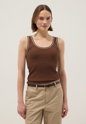 Giovane donna con capelli corti castani che indossa una canotta marrone con bordi bianchi e pantaloni beige con cintura nera, in piedi su uno sfondo chiaro.