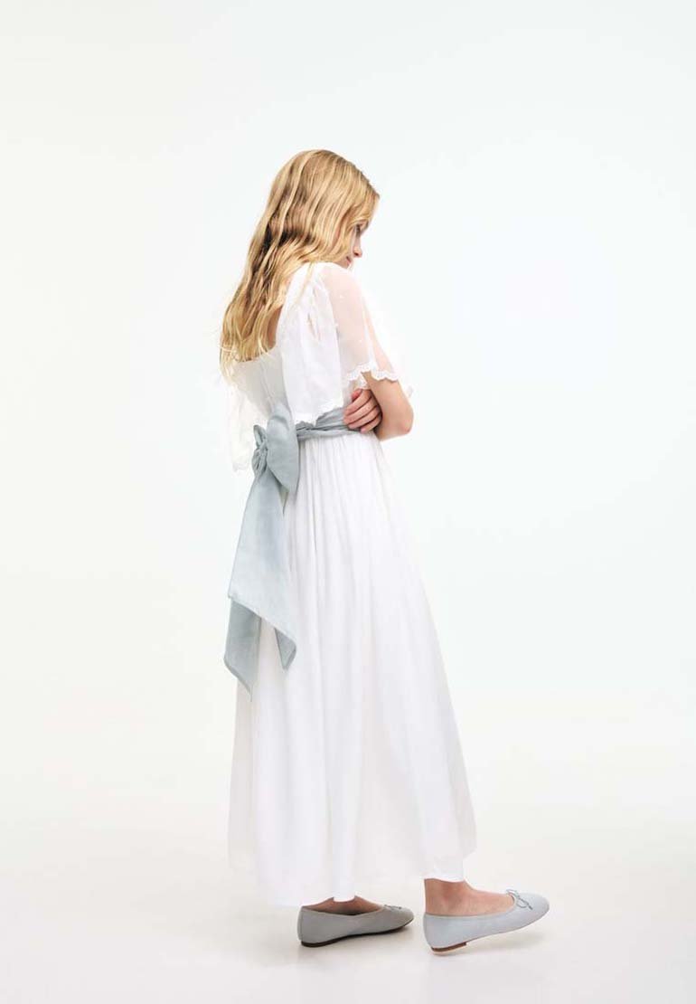Girl with long blonde hair wearing a white dress with sheer sleeves and a large light gray bow at the back, paired with gray ballet flats.