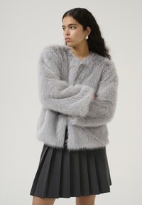 Giacca in finto pelo grigio chiaro con una superficie texturizzata e morbida. Presenta un collo rotondo e maniche lunghe, abbinata a una gonna plissettata scura.