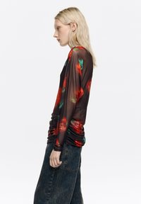 Bluza z długim rękawem z cienkiego czarnego materiału z żywym czerwono-zielonym wzorem kwiatowym. Posiada marszczone boki dla efektu marszczenia.