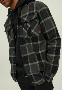 Giacca camicia a quadri nera e grigia realizzata in tessuto testurizzato, con due tasche sul petto, un accento rosso e chiusure a bottone sul davanti.