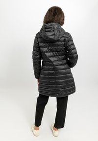 Zwarte pufferjas met een capuchon, voorzien van een horizontaal gestikt patroon, geribbelde manchetten en een knielange snit. Draagt over zwarte broeken en sneakers.