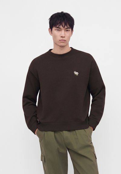 Jeune homme aux cheveux noirs courts portant un sweat-shirt marron foncé avec une petite broderie de fleur blanche et un pantalon cargo vert sur un fond uni.