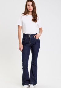 Ciemne dżinsowe spodnie dzwony mają wysoki pas z dwoma przednimi guzikami, kontrastowe przeszycia i kieszenie z przodu, w połączeniu z prostą białą koszulą.