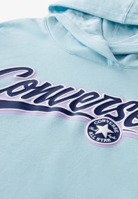 Ανοιχτό μπλε φούτερ με απαλή υφή, που διαθέτει μεγάλο, στυλιζαρισμένο λογότυπο «Converse» σε σκούρο μπλε με μοβ λεπτομέρειες, και έναν στρογγυλό λογότυπο από κάτω.