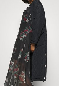 Den vadderade svarta långkoftan har tryckknappar och bärs över en blommig svart klänning med röda och gröna mönster, i halvtransparent tyg.