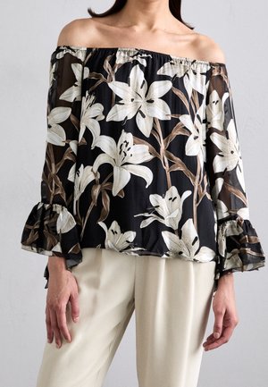 Kvinde iført sort off-shoulder bluse med hvidt blomstermønster og flæseærmer kombineret med lyse beige bukser.