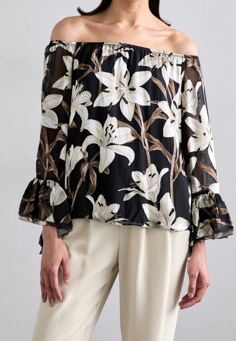 Femme portant un chemisier noir épaules dénudées avec un imprimé floral blanc et des manches cloche à volants, associé à un pantalon beige clair.