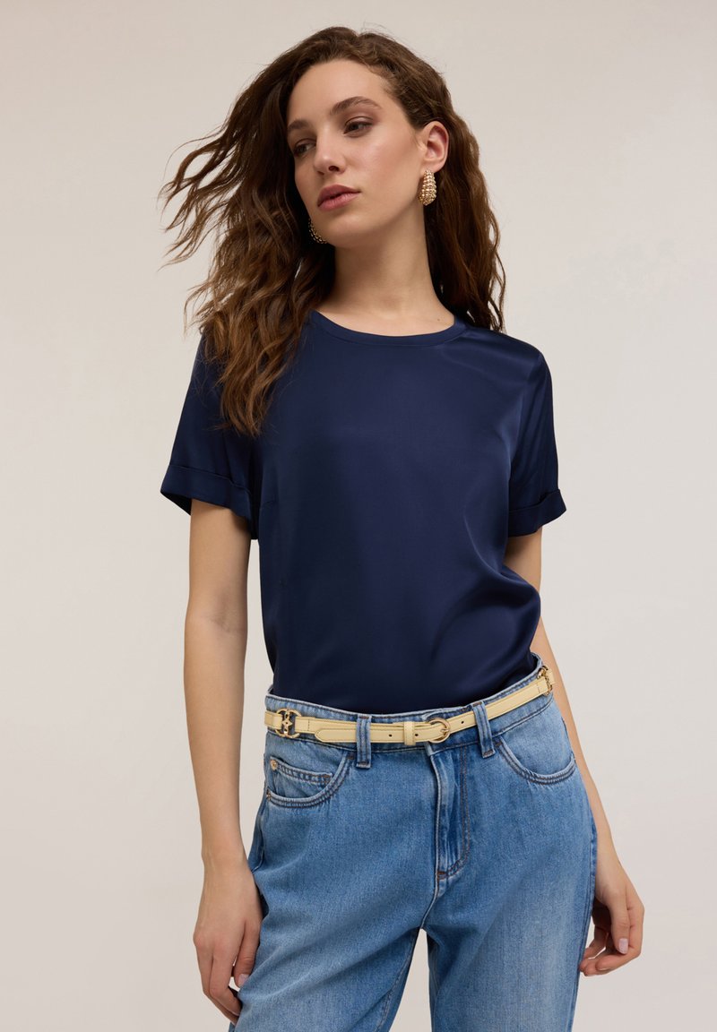 Blusa blu navy a maniche corte con una texture liscia, abbinata a jeans blu a vita alta e una cintura color crema con fibbia metallica.