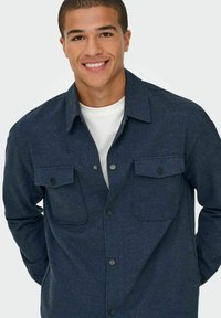 Camisa azul oscuro de botón con dos bolsillos frontales, botones negros y una tela texturizada. Lleva puesta una camisa de color claro. Diseño de cuello sencillo.