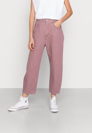 Henkilö, joka pukeutunut vaaleanpunaisiin löysiin korkeavyötäröisiin farkkuihin, valkoiseen hihattomaan toppiin ja valkoisiin korkeavartisiin tennareihin, seisoo yhdellä kädellä taskussa.