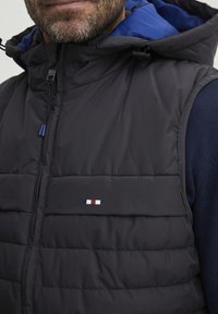 Prsluk s punjenjem u tamnoplavoj boji, s kapuljačom i plavim unutarnjim oblogom. Horizontalni prešani uzorak s malim, višebojnom logotipom na prsima.
