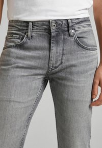 Uomo che indossa jeans in denim grigio sbiadito attillati e una t-shirt bianca infilata dentro, con tasche frontali e chiusura a bottone.
