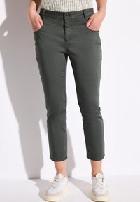 Dunkelgrüne cropped Hosen mit zwei Seitentaschen, taillierter Passform, glatter Textur und ergänzt durch weiße Sneakers.