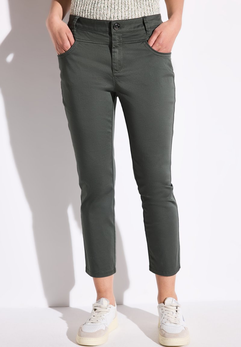 Dunkelgrüne cropped Hosen mit zwei Seitentaschen, taillierter Passform, glatter Textur und ergänzt durch weiße Sneakers.