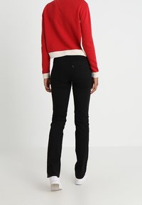 Osoba w czerwono-białym swetrze, czarnych dopasowanych spodniach i białych trampkach, stojąca na jednolitym jasnym tle, zwrócona plecami.