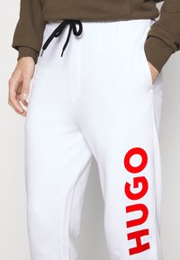 Hvide sweatpants med en sort snor og sidelommer. Har et stort rødt "HUGO" logo trykt lodret på venstre ben.
