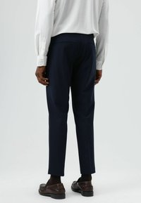 Marineblaue Hosen mit schmaler Passform. Hergestellt aus glattem Stoff, mit zwei Gesäßtaschen. Kombiniert mit einem weißen Langarmshirt und dunklen Schuhen.