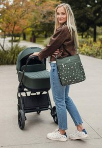 Zelený kočárek s hladkým, zaobleným designem; zelená taška přes rameno s květinovým vzorem; žena v hnědé kordové bundě a modrých džínách.