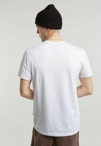 T-shirt in cotone bianco con collo rotondo, indossata sotto un berretto a coste nero. La schiena del modello è visibile, evidenziando il design semplice e la morbidezza del tessuto.