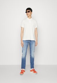 Polo shirt blanc avec col, jeans bleu clair et baskets en daim orange, accessoirisé avec des lunettes de soleil noires. Design simple, coupe décontractée.