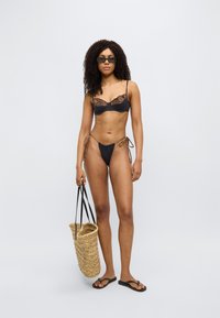 Μαύρο μπικίνι με λεπτομέρειες δαντέλας, σλιπ χαμηλής μέσης και δεσίματα στο πλάι. Το μοντέλο κρατάει πλεκτή ψάθινη τσάντα με μαύρα λουριά. Σανδάλια με επίπεδο πάτο.