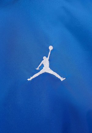 Niebieska tkanina o gładkiej fakturze z białym haftowanym logo Jumpman, przedstawiającym sylwetkę postaci w powietrzu z piłką do koszykówki.