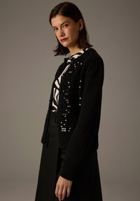 Schwarze Strickjacke mit Paillettenverzierungen, strukturiertem Stoff und Knopfverschluss, kombiniert mit einem schwarz-weißen gemusterten Oberteil.