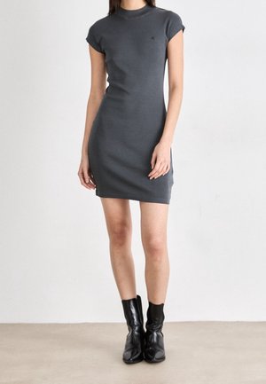 Woman wearing a short-sleeve, dark gray bodycon dress paired with black ankle boots, standing against a plain light background.