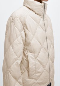 Beżowa pikowana kurtka z wysokim kołnierzem, przednim zamkiem błyskawicznym i boczną kieszenią na zamek, noszona przez osobę, której zdjęcie jest przycięte od ust do talii.