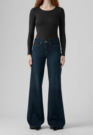 Femme portant un jean bleu foncé à jambes larges et un top noir ajusté à manches longues, debout devant un fond clair uni.