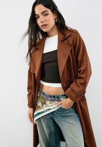 Hnědý zamšový trenčkot s detaily na knoflíky, přehozený přes hnědý crop top a vzorované džíny, s multicolor akcenty a třepenými okraji.