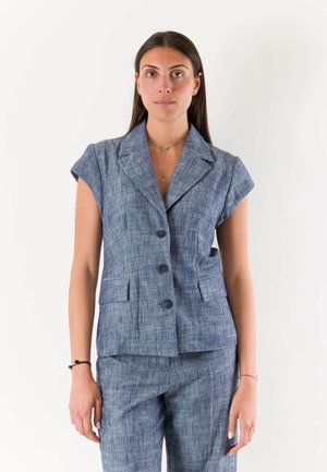 Donna che indossa un blazer blu strutturato senza maniche con pantaloni abbinati, in piedi su uno sfondo bianco semplice.