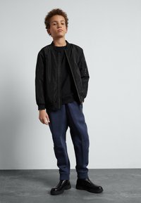 Schwarze Bomberjacke mit gerippten Bündchen, getragen über einem schwarzen Sweatshirt. Dunkelblaue Jogginghosen und schwarze Plateau-Stiefel vervollständigen das Outfit.